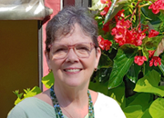 A woman with short hair and glasses, wearing a light green top and beaded necklaces, stands smiling in front of a lush green plant with red flowers